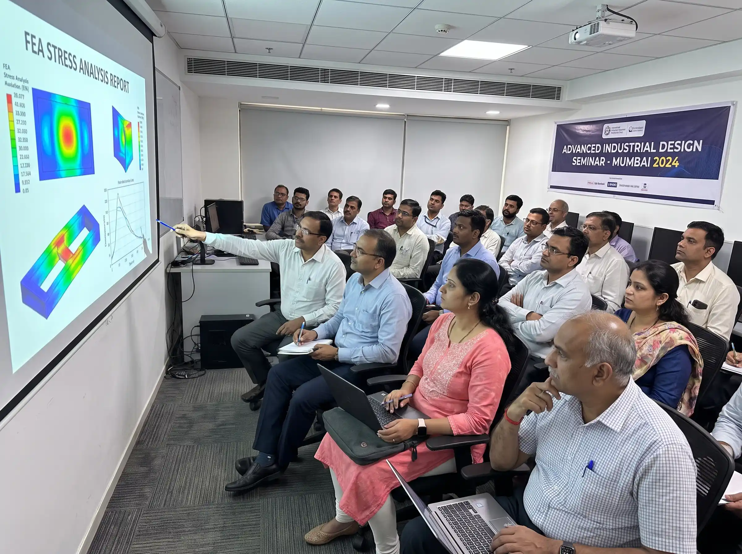 A group of focused Indian engineering professionals attending an advanced industrial design seminar, looking at a projector displaying stress analysis data.